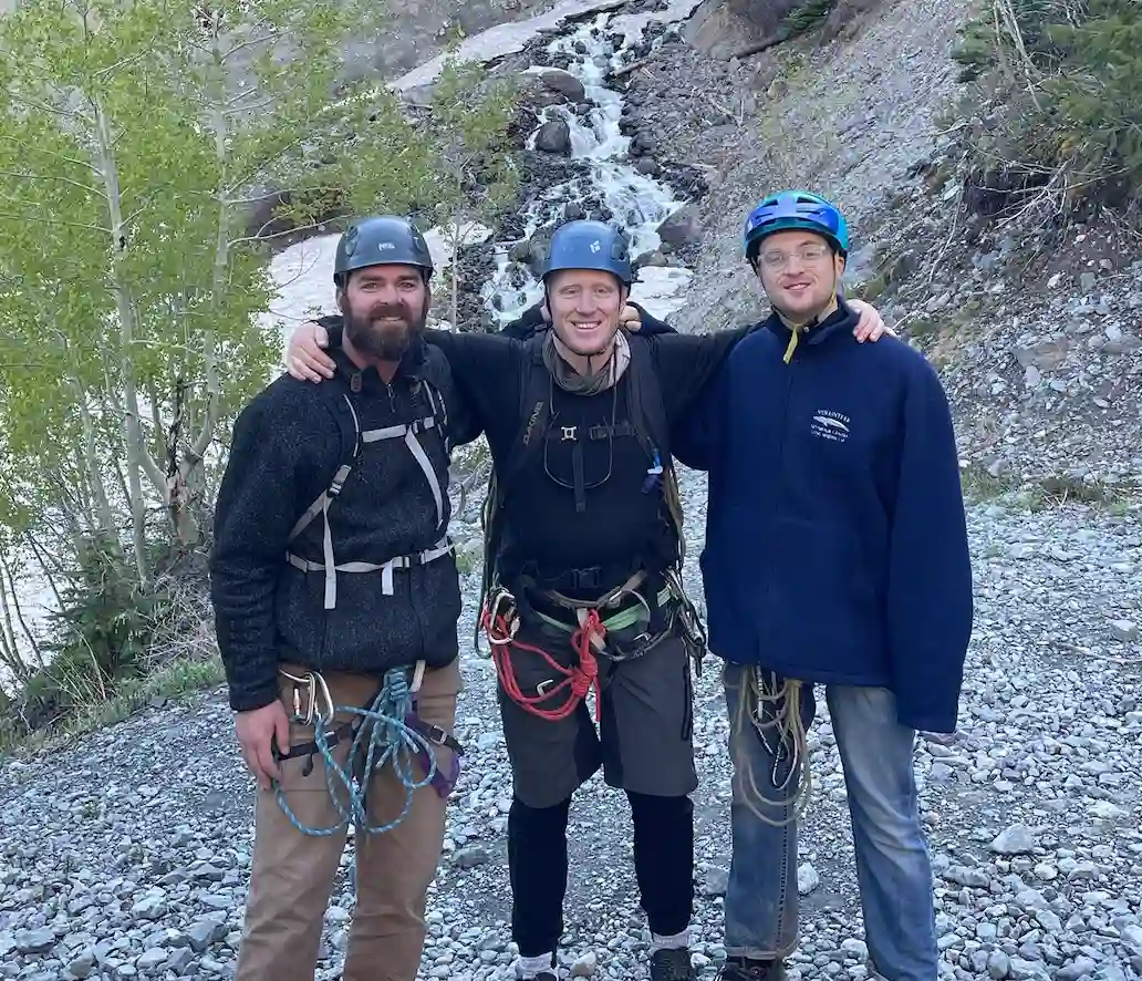 Three people posing outdoors with climbing gear in the mountains
